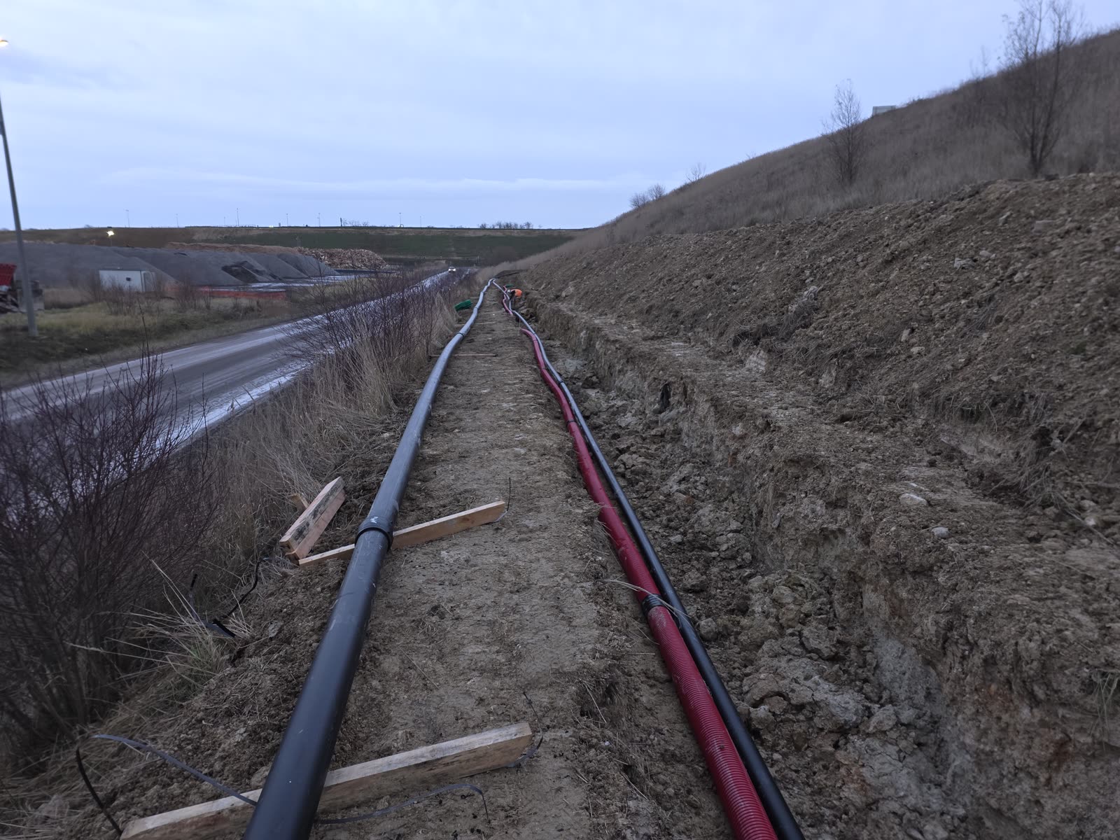 Tranchée de réseaux ouverte le long d'une route de chantier, en fin de journée, deux fourreaux posés en fond de fouille